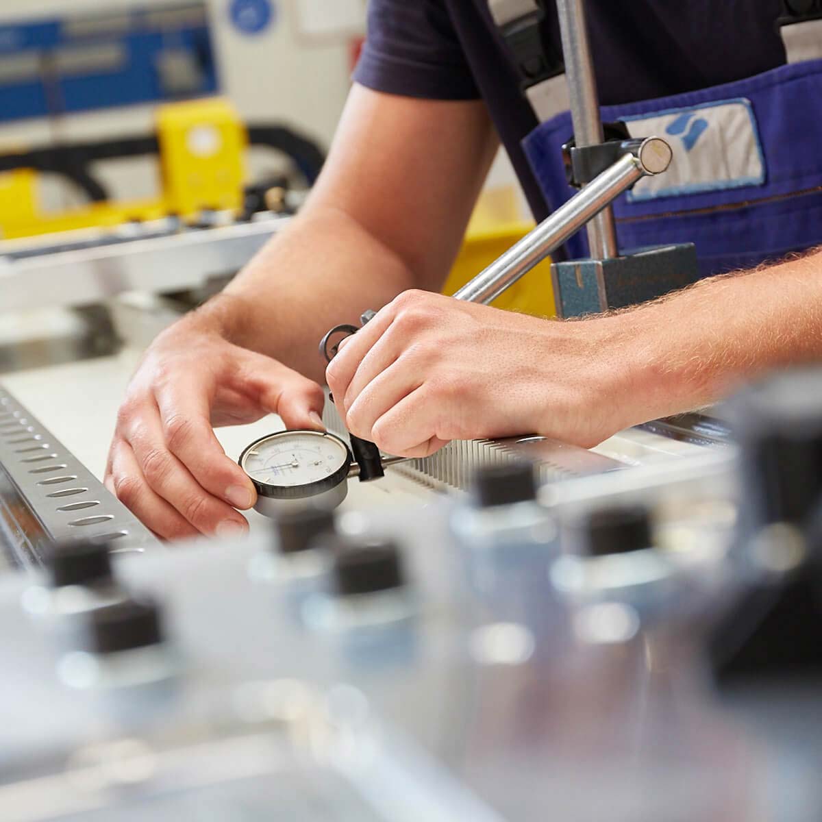 Una persona sta usando un comparatore a quadrante per misurare un oggetto in un'officina, circondata da strumenti e attrezzature.
